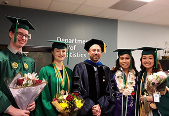 A group of students in green caps and gowns posing with their professor on graduation day. 